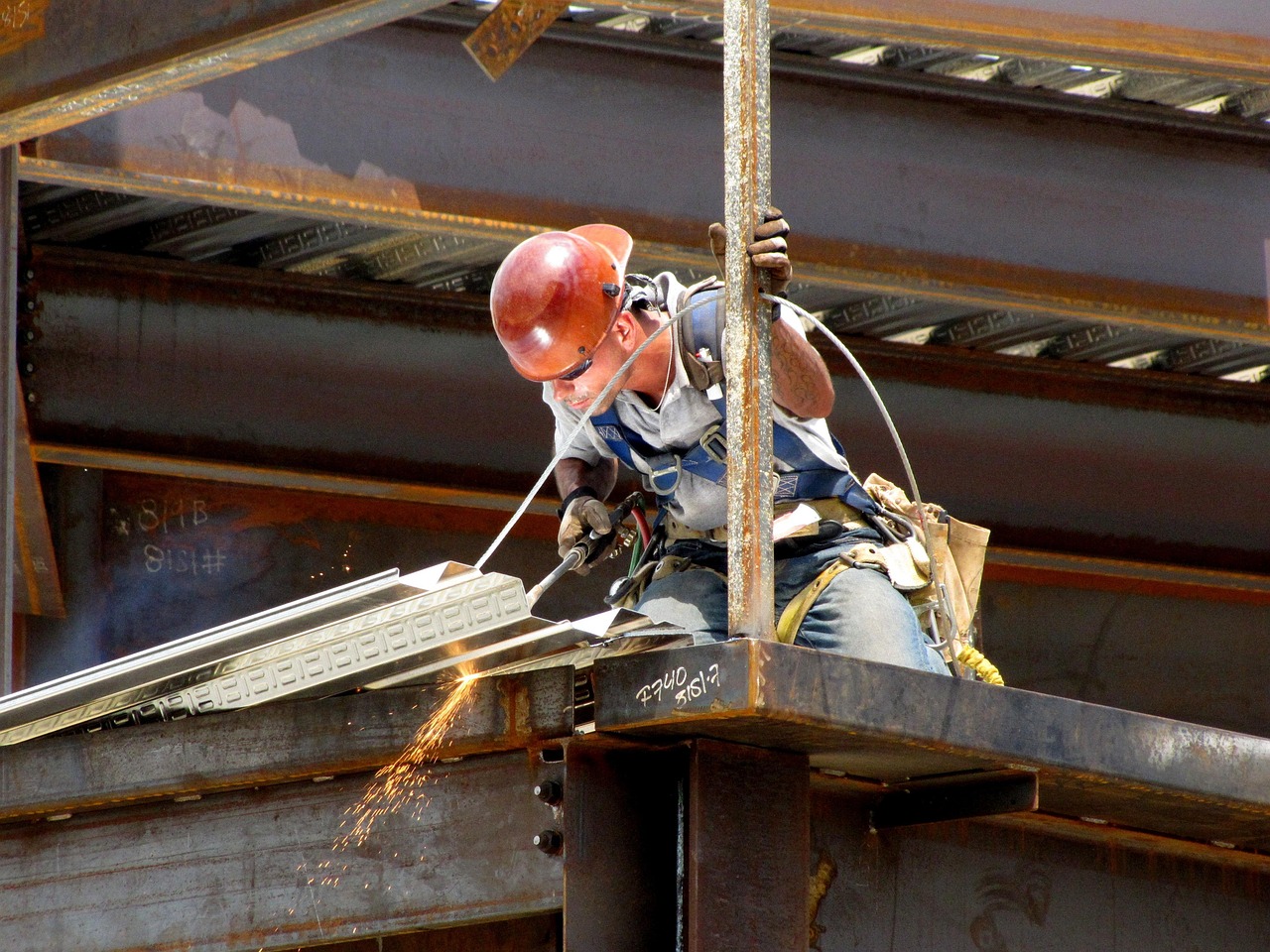 Construction worker wearing safety gear and helmet welding steel beam support systems on a building structure, with sparks flying during the process.